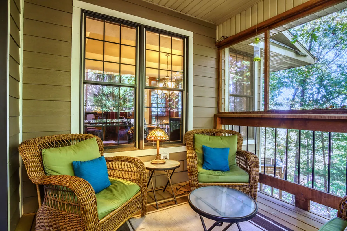 A cozy breakfast nook with wicker chairs, decorative cushions, a small table, and a lamp. Windows surround the space, filling it with gentle morning light and offering calming views of the outdoors—a perfect spot for casual coffee or intimate morning chats.