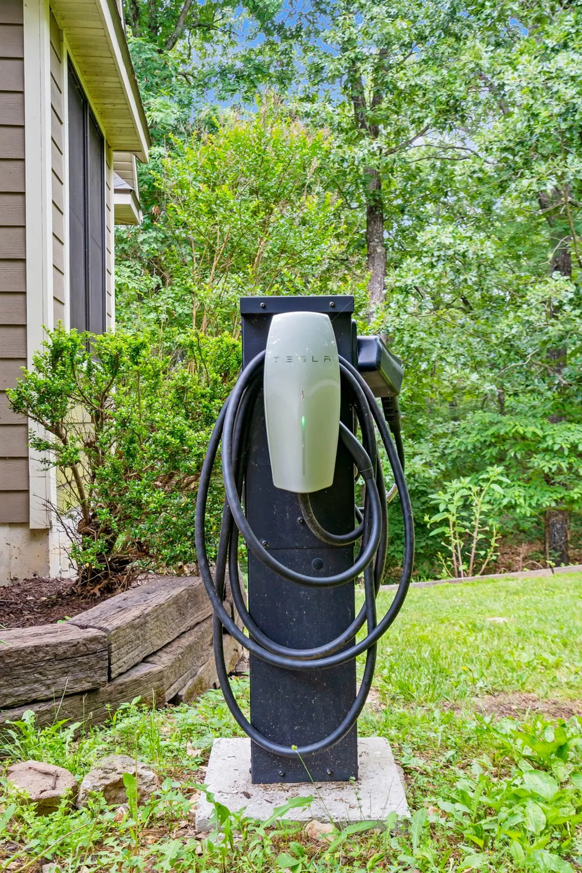This exterior image showcases a Tesla charging station prominently placed beside the house, with lush greenery surrounding the area. The exterior wall of the home is visible, grounding the charging feature in the estate’s natural landscape.