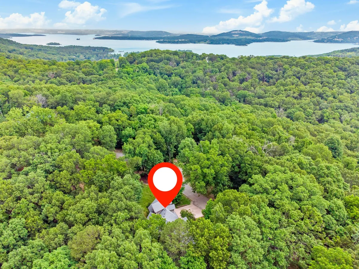 An inviting exterior view of the house nestled in lush greenery, with Table Rock Lake visible in the background and a winding driveway leading to the property. The scene evokes a sense of peaceful seclusion, natural beauty, and immediate proximity to lakeside adventures.