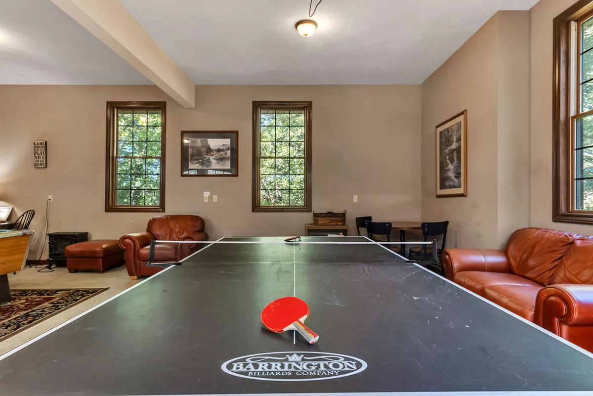 A playful and welcoming space with a prominent ping-pong table, leather couches, large windows with a view to greenery, and colorful artwork on the walls. The room encourages both active play and relaxed socializing, ideal for game nights or tournaments.