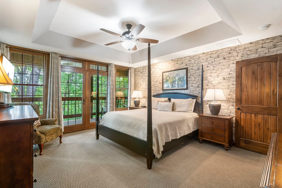 A serene bedroom featuring a four-poster bed, stone accent wall, large windows letting in natural light, and wooden furniture. The decor is tranquil with tasteful New England-inspired touches, and the room looks spacious and welcoming.
