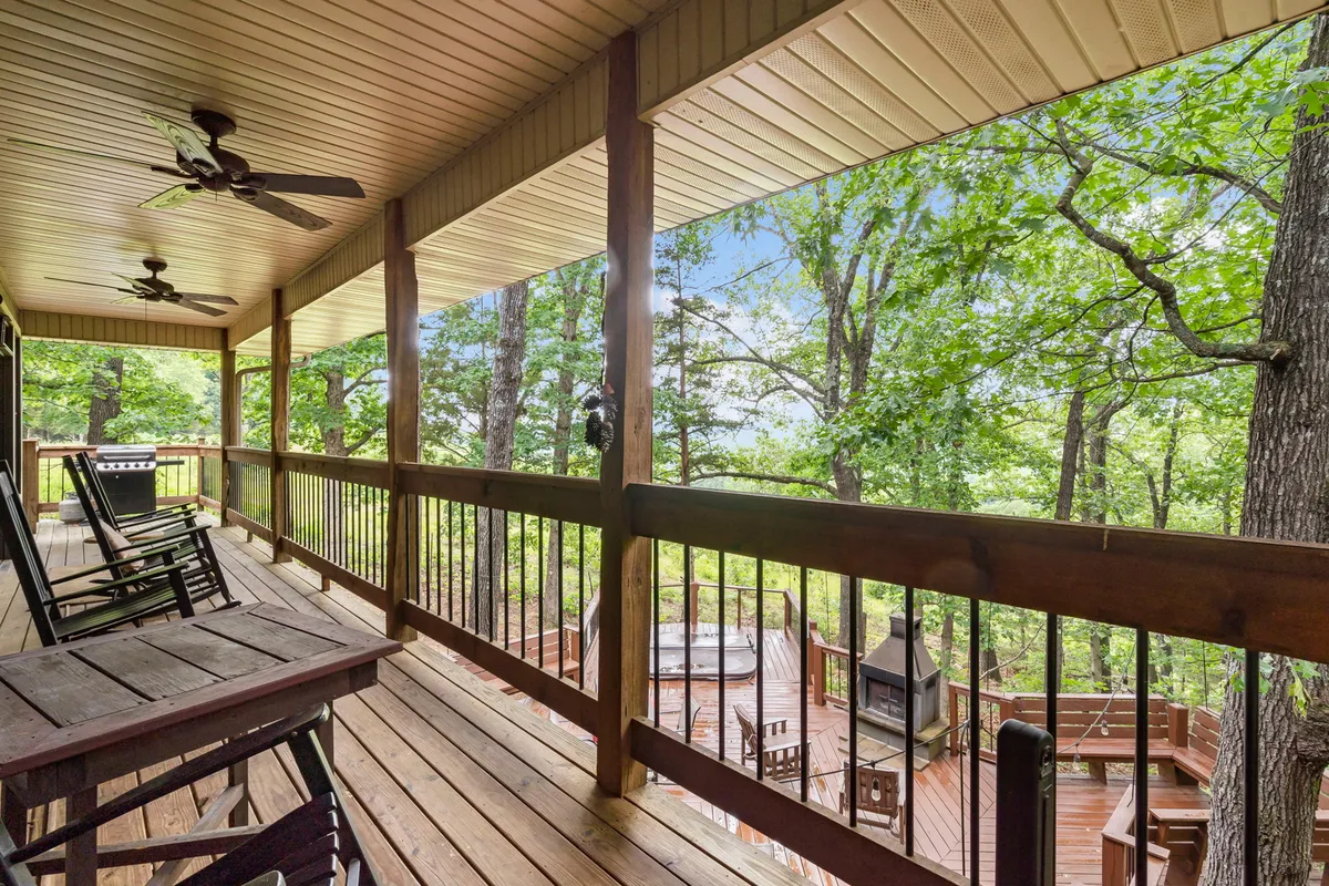 An exterior shot featuring rocking chairs, a wooden railing, surrounding trees, an outdoor grill, and a visible hot tub area. This area is set up for relaxation, socializing, and enjoying nature with loved ones.