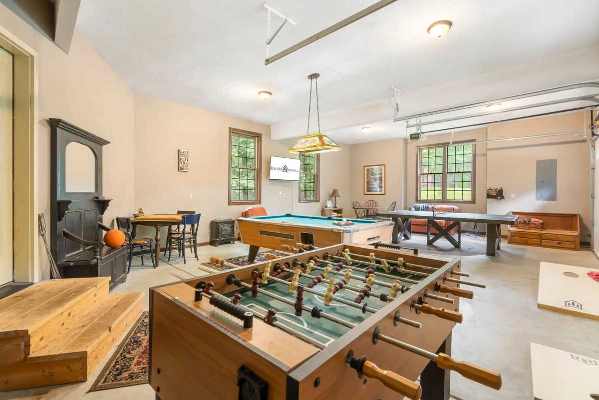 A lively interior scene highlighting both a foosball table and a pool table, surrounded by wooden chairs and illuminated by natural light streaming through large windows. The mood is casual and fun, set up for social games or celebrations.