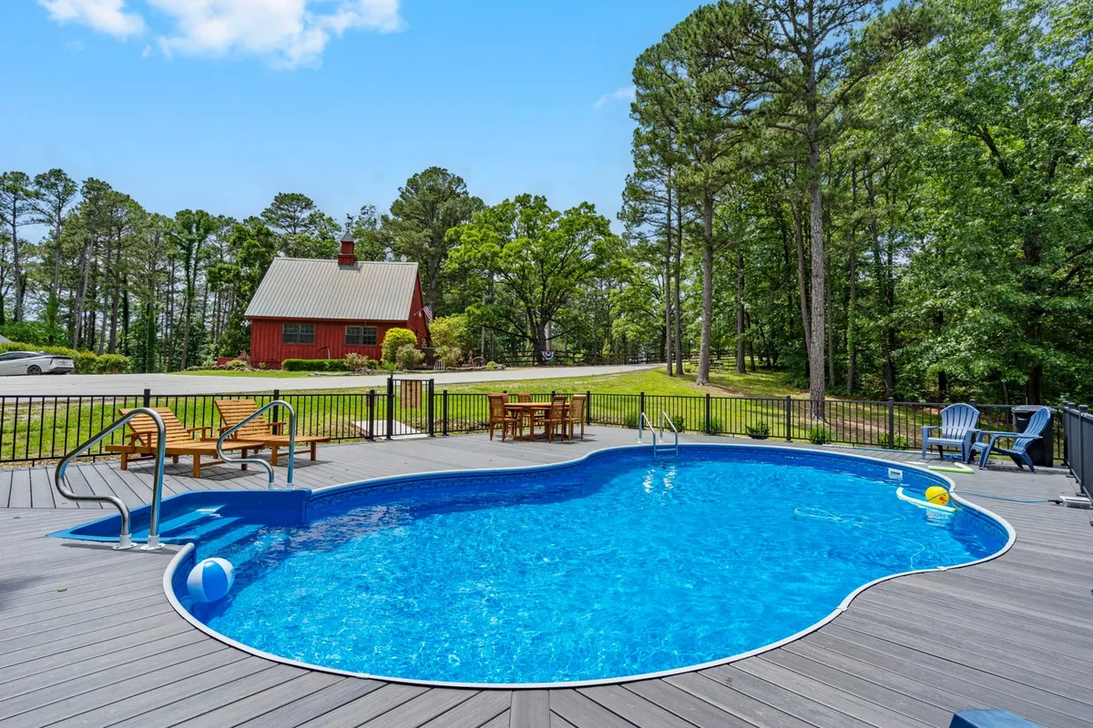 A sparkling swimming pool adjacent to a red house with a wooden deck and lounge chairs, set amid greenery. The scene is perfect for sunbathing, swimming, or relaxing outdoors, and reflects the shared estate amenities available just beyond the private decks.