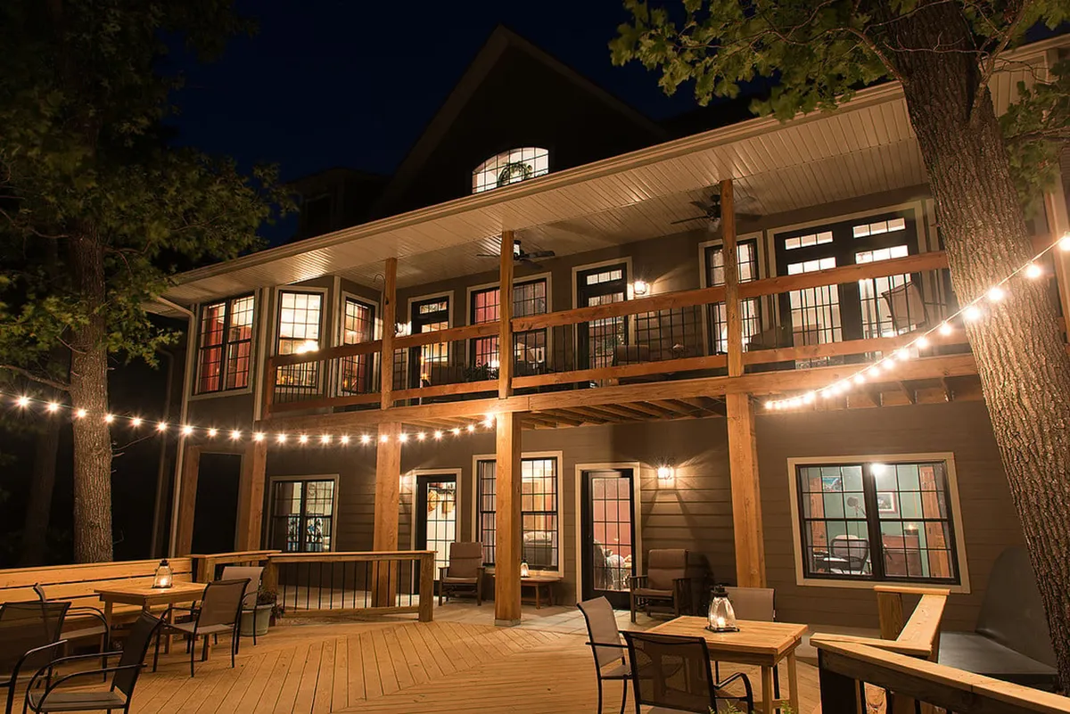 The hero image shows the exterior of Meadow's Edge Lodge at dusk, with inviting string lights illuminating a spacious wooden deck and balcony. Warm light spills from the windows, creating a cozy glow that hints at lively gatherings inside. The setting overlooks a serene, tree-lined meadow, perfect for outdoor dining, evening relaxation, and celebrating under the stars.