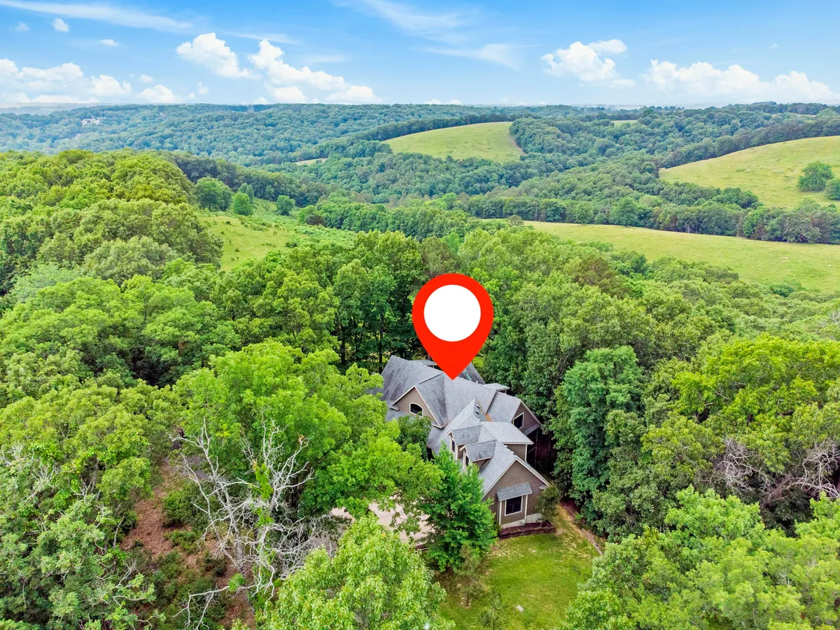 This image displays the house nestled among mature trees and rolling hills, with a wide view of the property’s peaceful natural surroundings. The inviting porch and open landscape capture the essence of rural tranquility.