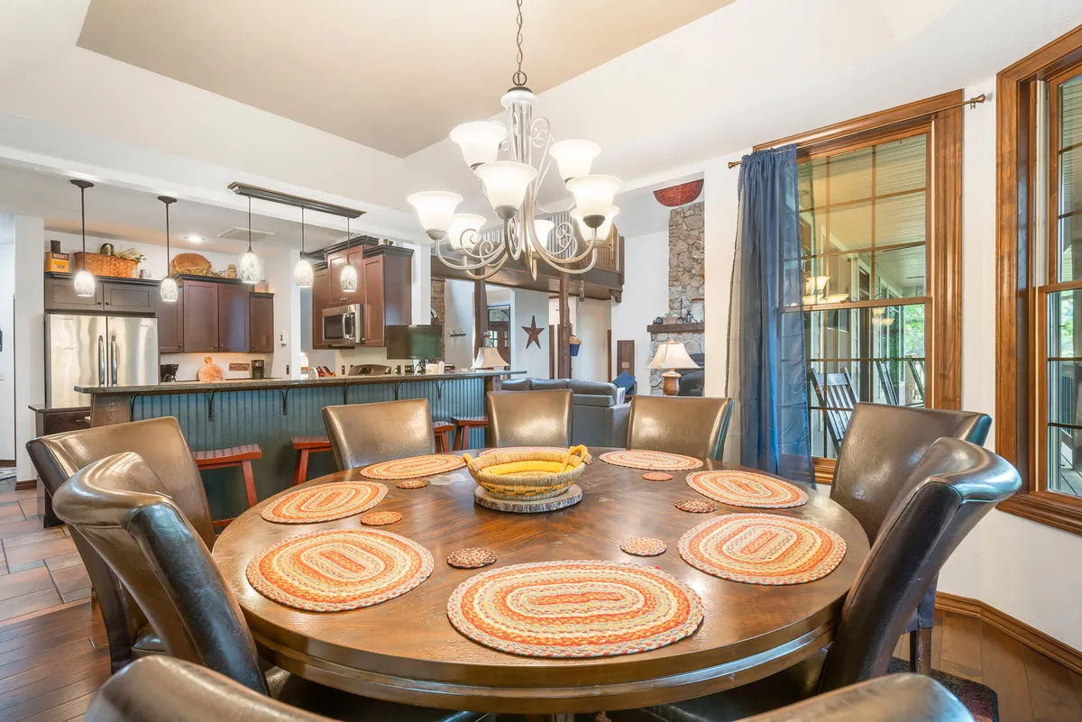 A circular dining room with a round wooden dining table set beneath an elegant chandelier. The area is surrounded by large windows that invite in natural light and reveal serene outdoor views. The kitchen is visible in the background, illustrating the seamless flow between the dining and cooking spaces.