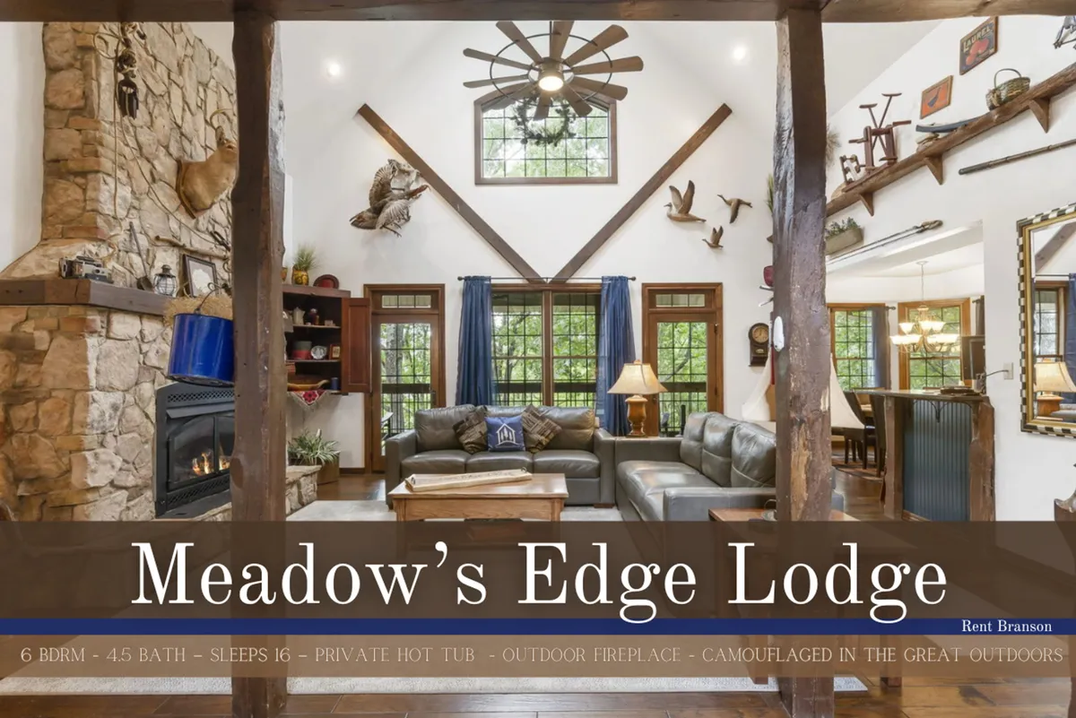 A spacious great room featuring a majestic stone fireplace framed by large windows that flood the space with natural light. Hand-hewn wooden beams arch overhead, and plush seating is arranged to encourage conversation and relaxation. The view through the windows reveals tranquil meadow scenery, connecting the indoors with the outdoors.